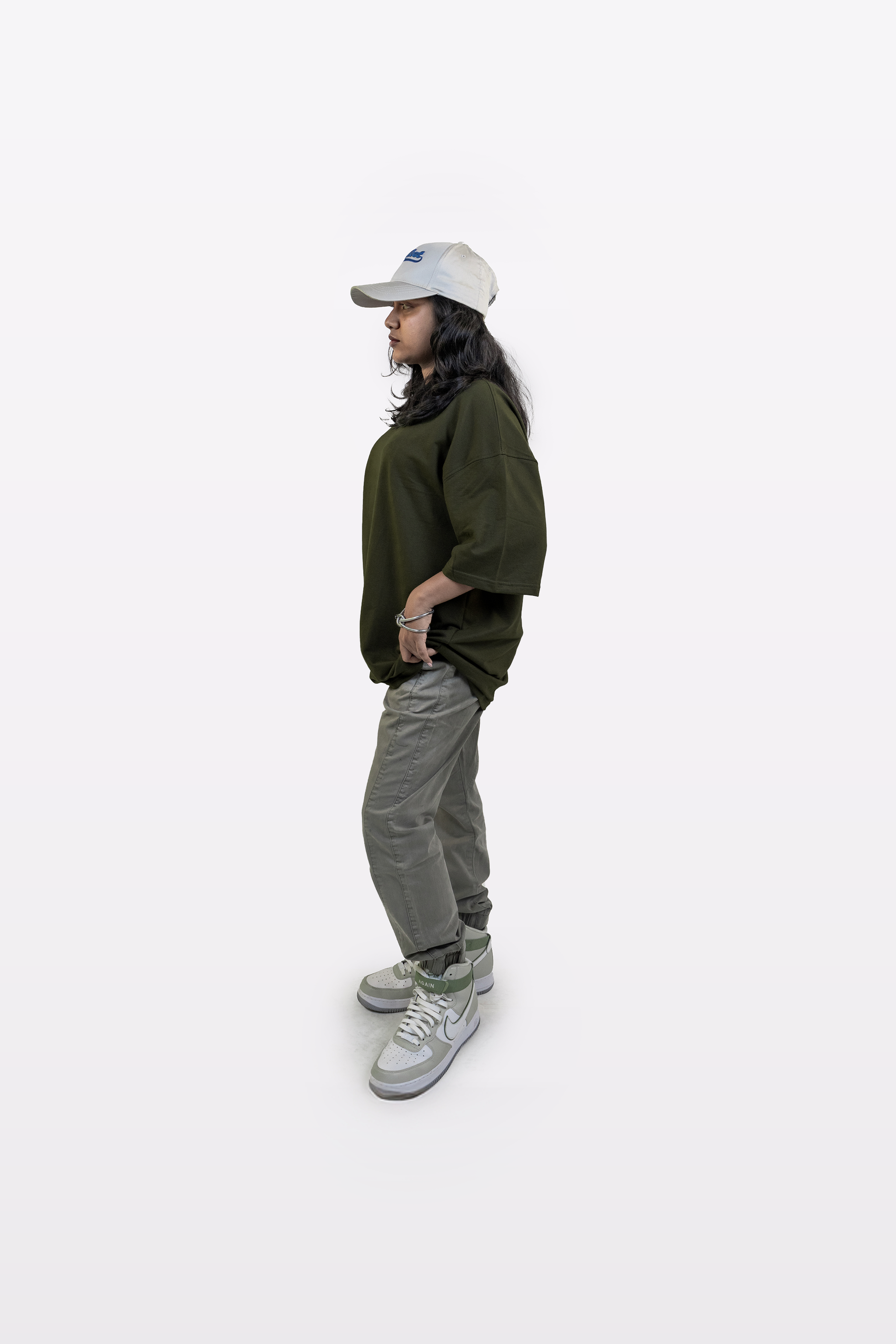 Side profile of a model wearing the Harper Slein oversized t-shirt in Nori Seaweed Green, paired with gray pants, white sneakers, and a white cap, standing against a white background.
