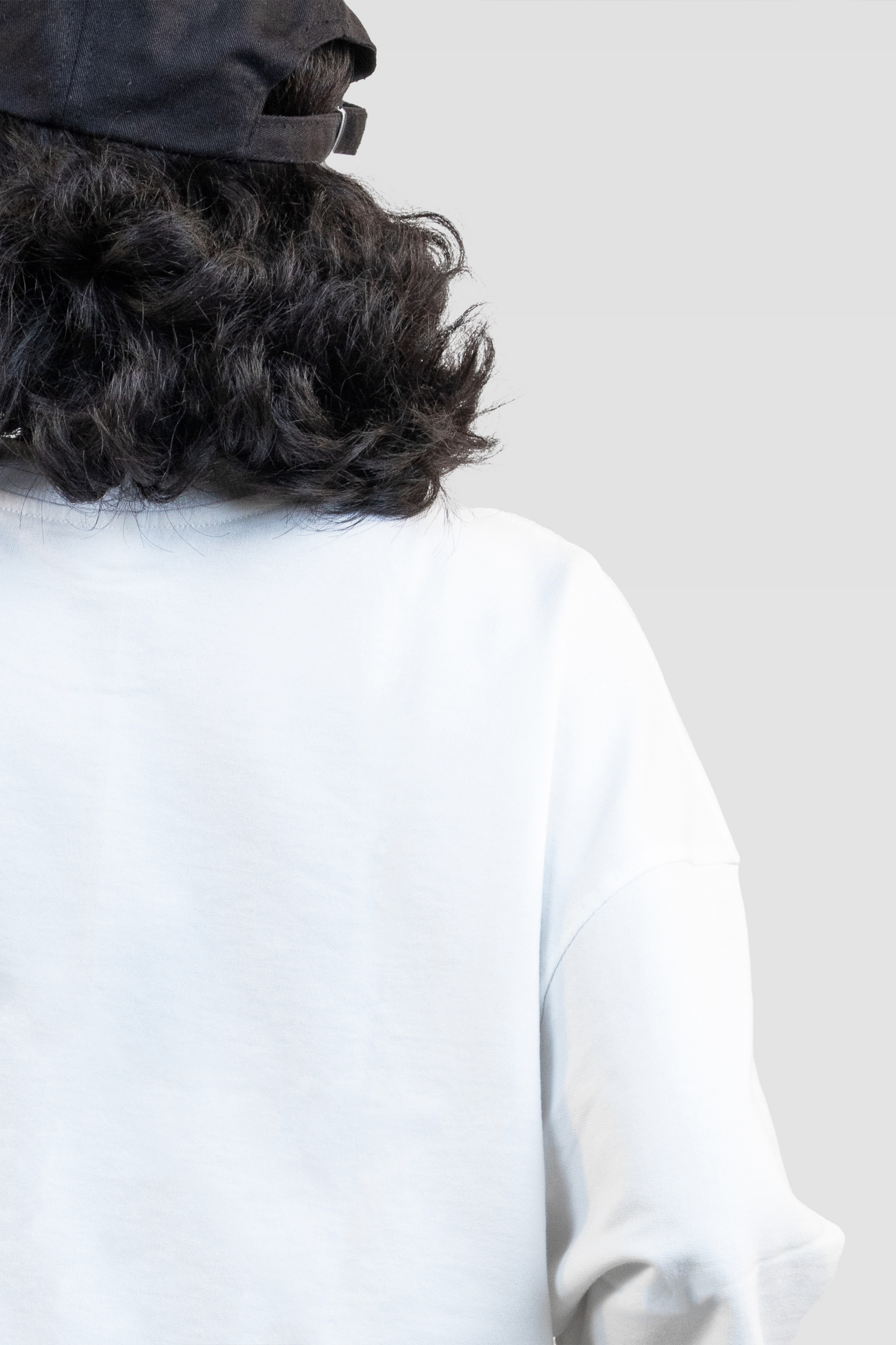 Back close-up of a female model wearing Harper Slein oversized t-shirt in Blank White with a black cap against a light gray background