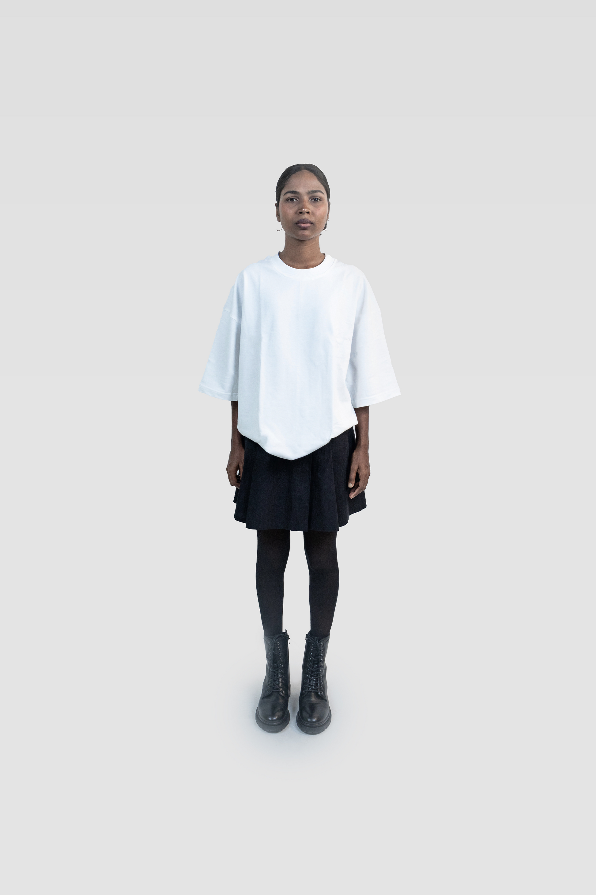 Front full-body view of a female model wearing Harper Slein oversized t-shirt in Blank White, styled with a black skirt and boots on a light gray background