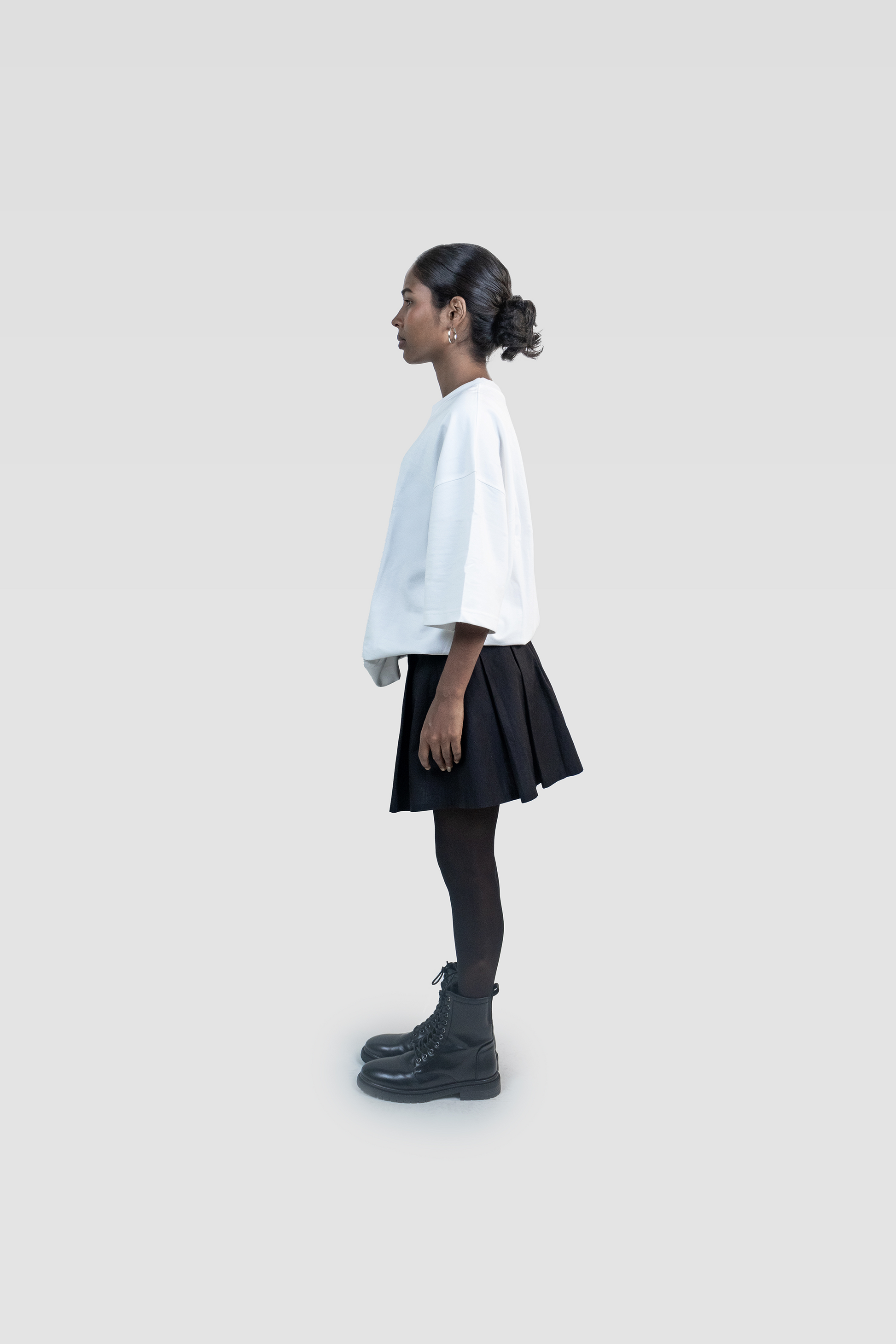 Side profile of a female model wearing Harper Slein oversized t-shirt in Blank White, styled with a black skirt and boots on a light gray background