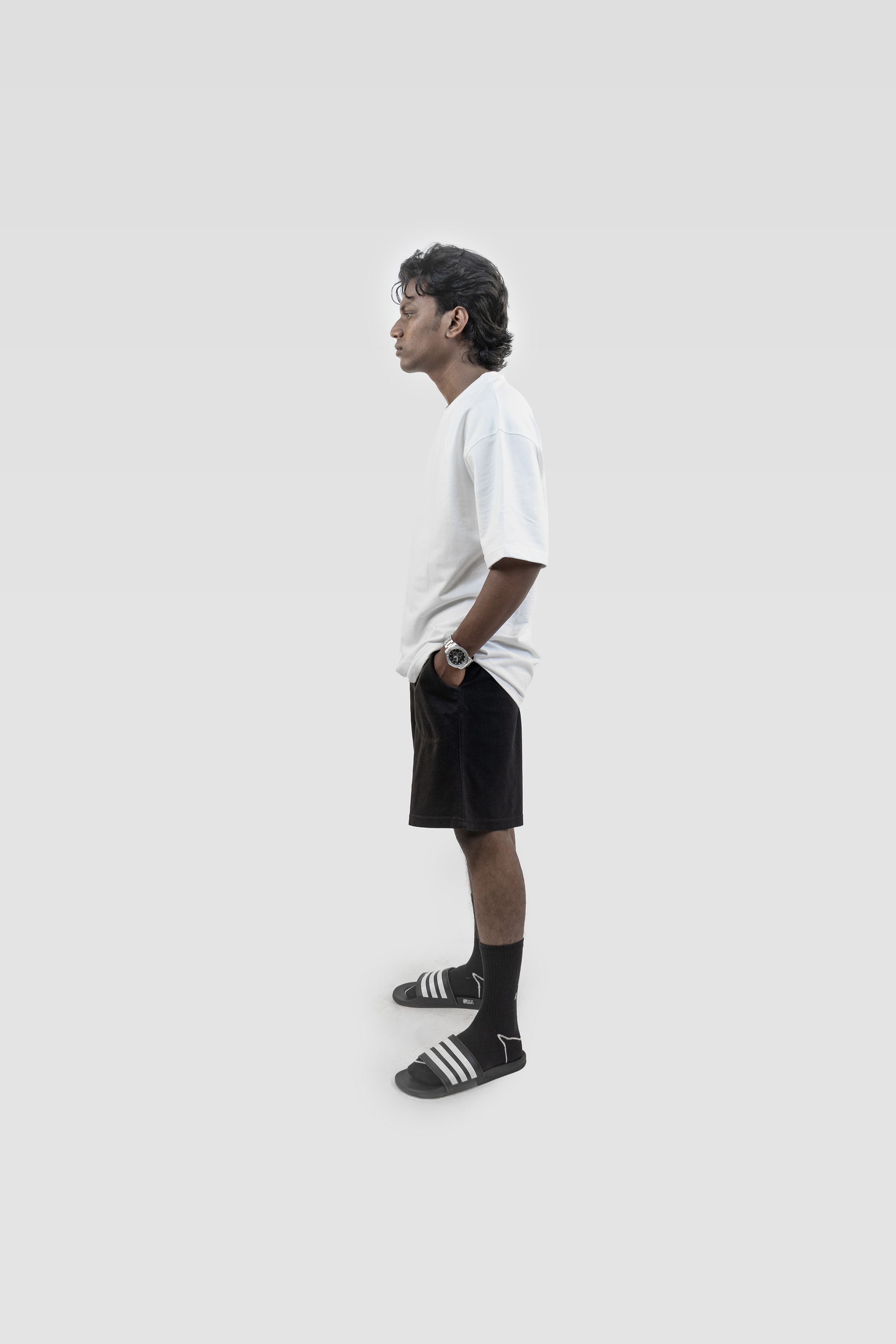 Side profile of a model wearing Harper Slein oversized t-shirt in Blank White, paired with black shorts and striped slides, against a light background