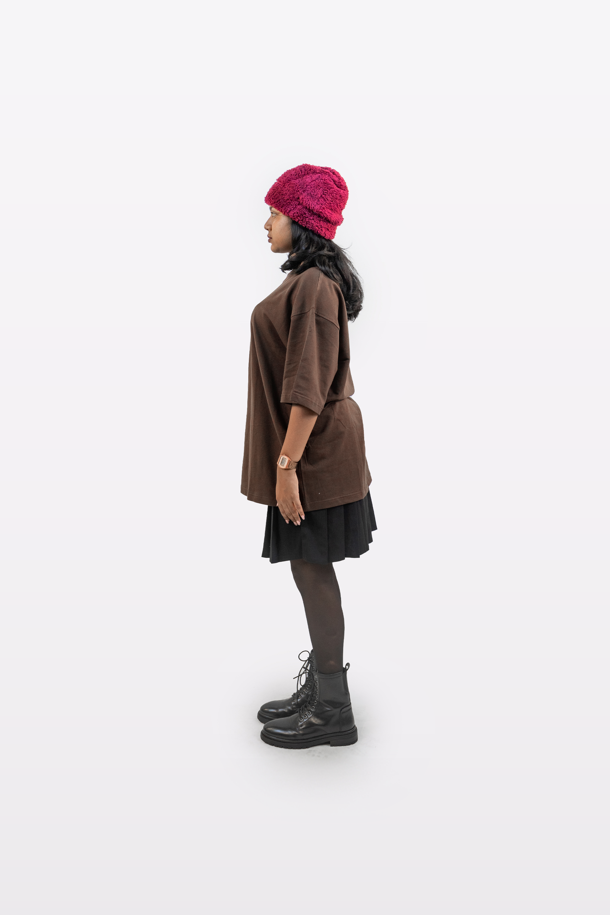 Side profile of a female model wearing Harper Slein oversized t-shirt in Cacao styled with a black skirt, black boots, and a red beanie on a white background