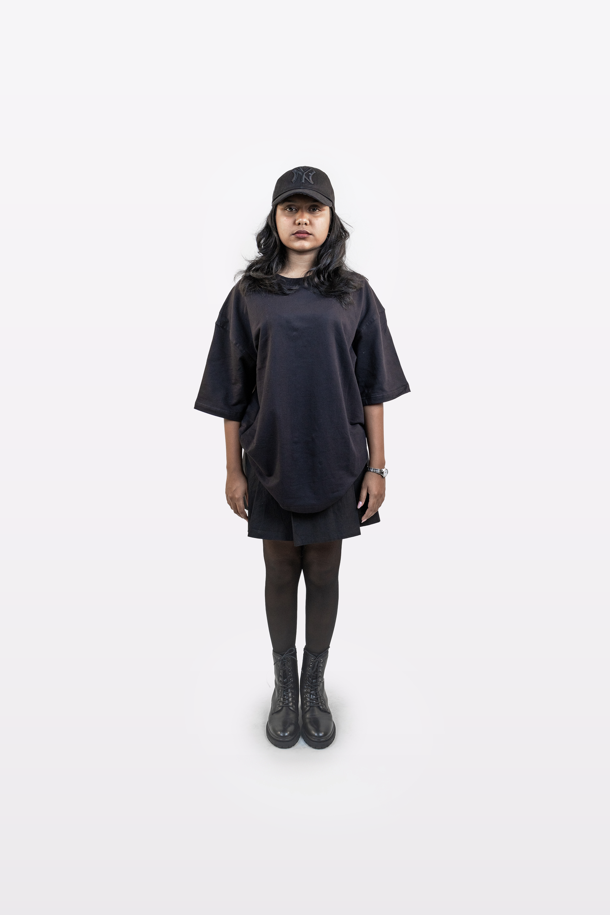 Front-facing full-body view of female model wearing Harper Slein oversized t-shirt in Classic Black, styled with black cap, skirt, tights, and boots against a white background