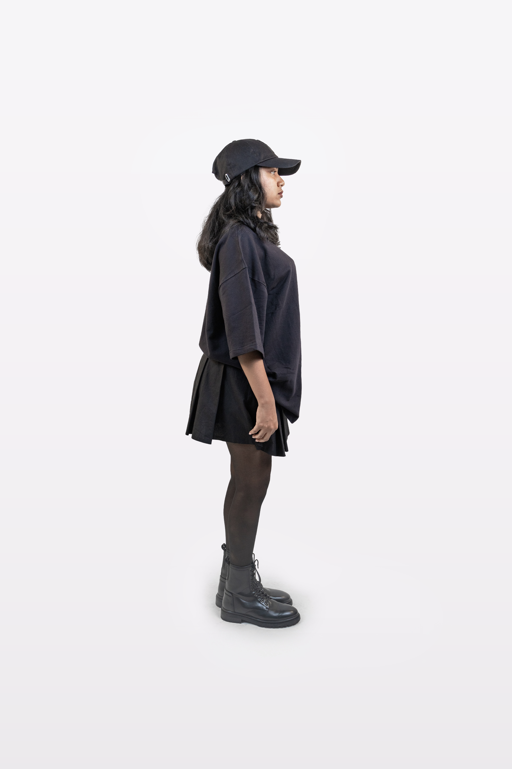 Side profile view of female model wearing Harper Slein oversized t-shirt in Classic Black, styled with black cap, skirt, tights, and boots on a white background