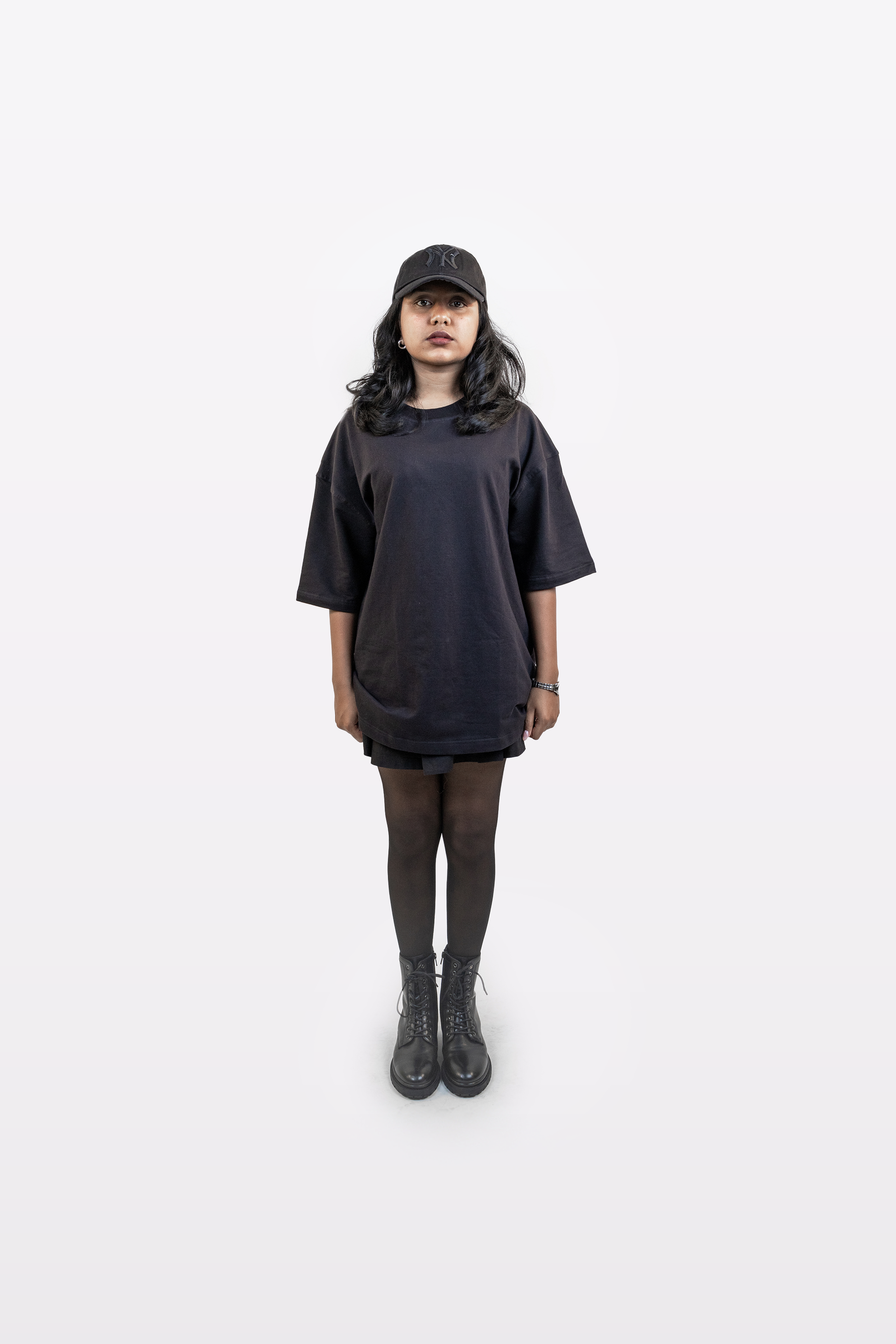 Full-body view of female model wearing Harper Slein oversized t-shirt in Classic Black, styled with black cap, tights, and boots on a white background