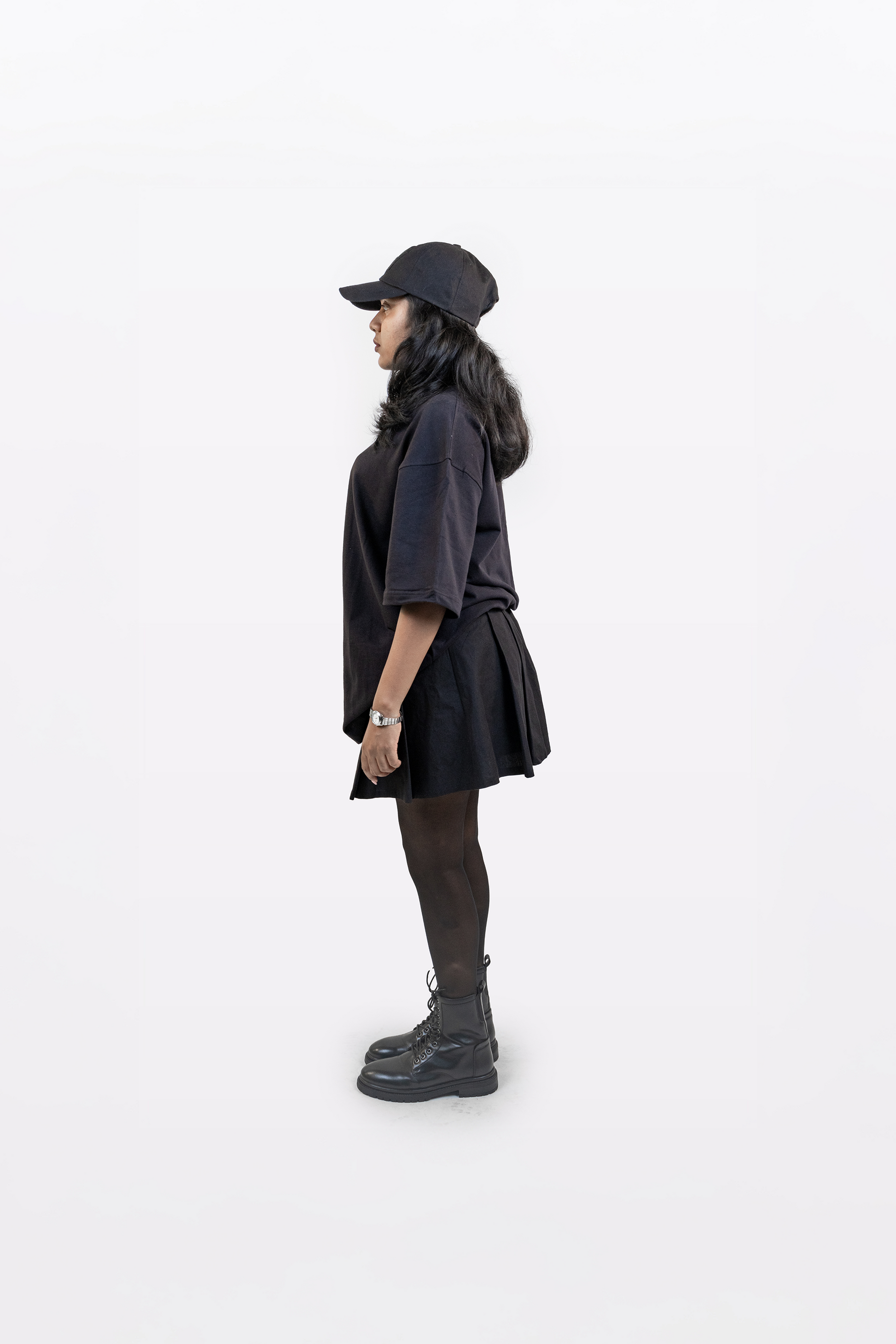 Upright side profile view of female model wearing Harper Slein oversized t-shirt in Classic Black, styled with black cap, skirt, tights, and boots on a white background