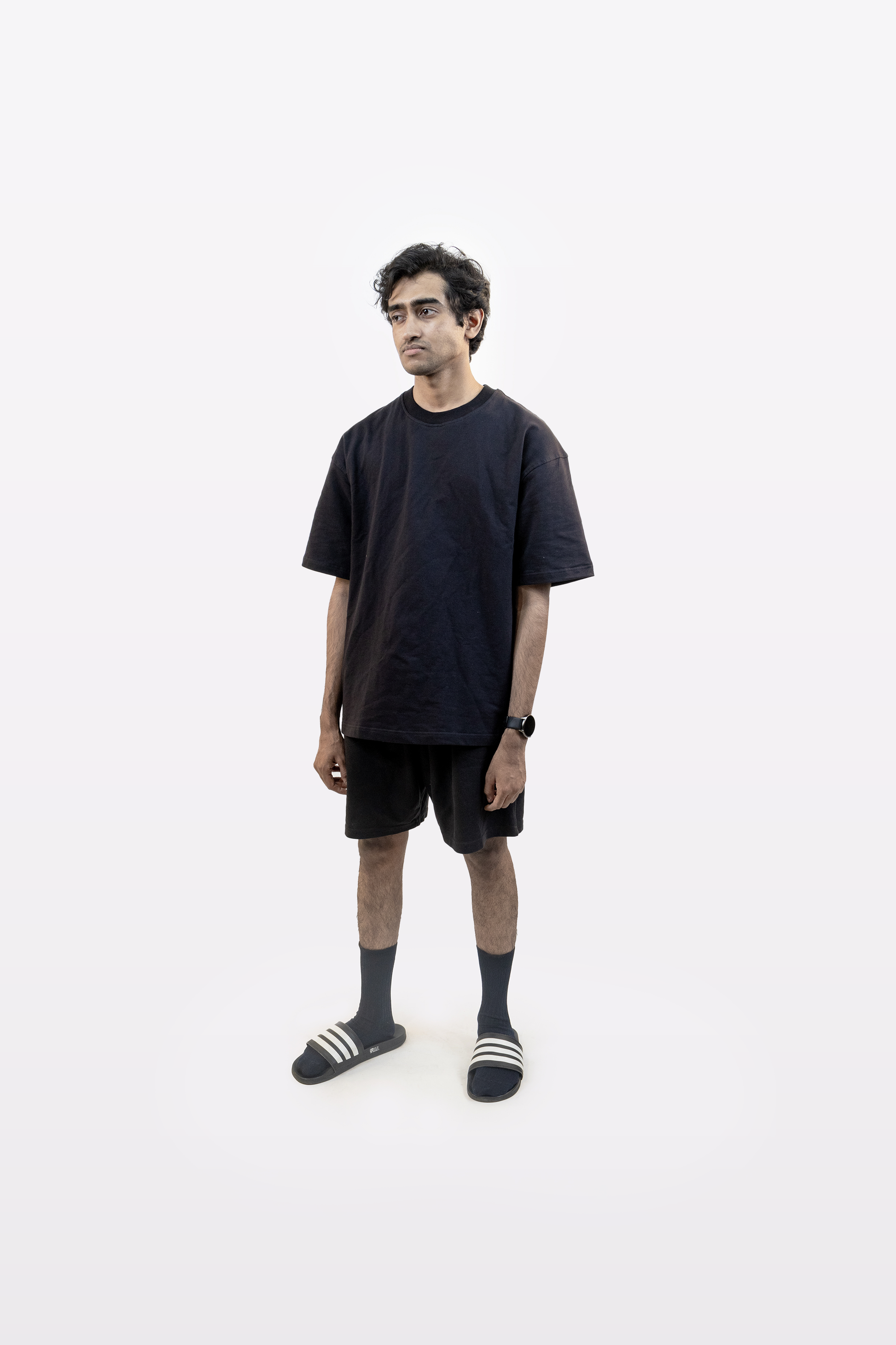Male model in full body front view wearing Harper Slein oversized t-shirt in Classic Black with black shorts, socks, and slides on a white background