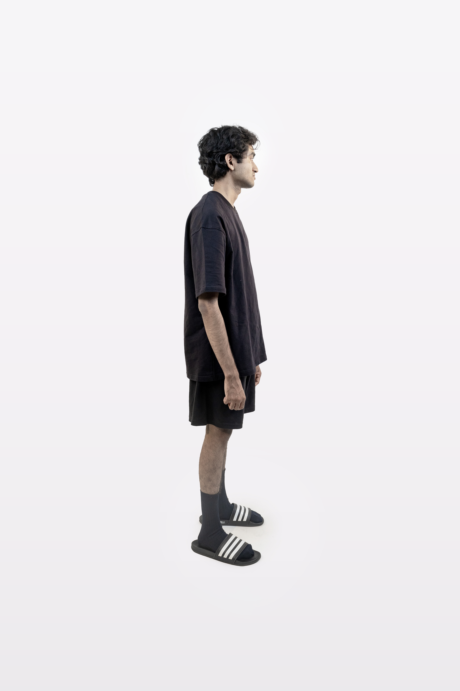 Male model in full body side profile wearing Harper Slein oversized t-shirt in Classic Black with black shorts, socks, and slides on a white background