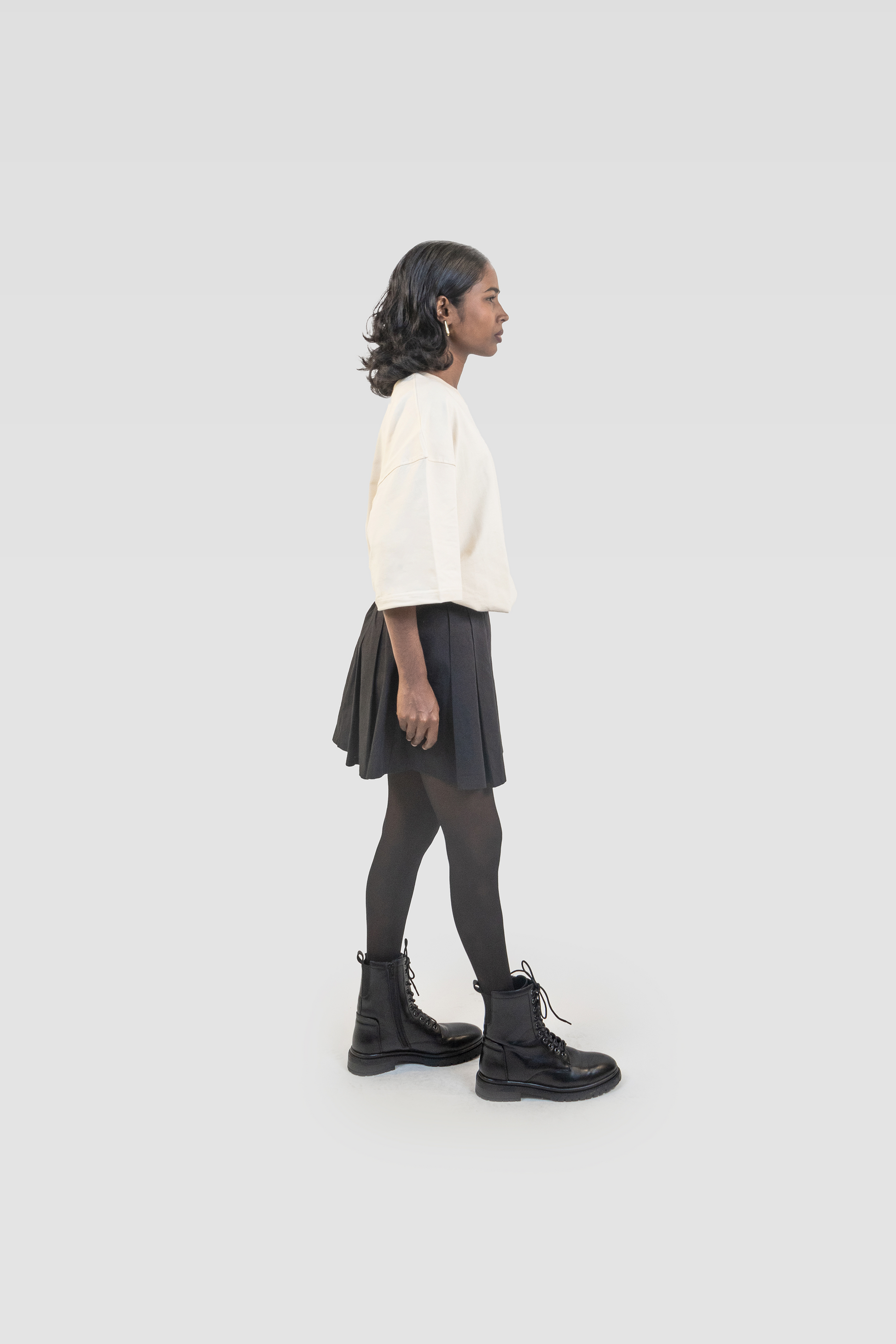 Side view of model wearing Harper Slein oversized t-shirt in OG Off White styled with black skirt, tights, and boots on a white background