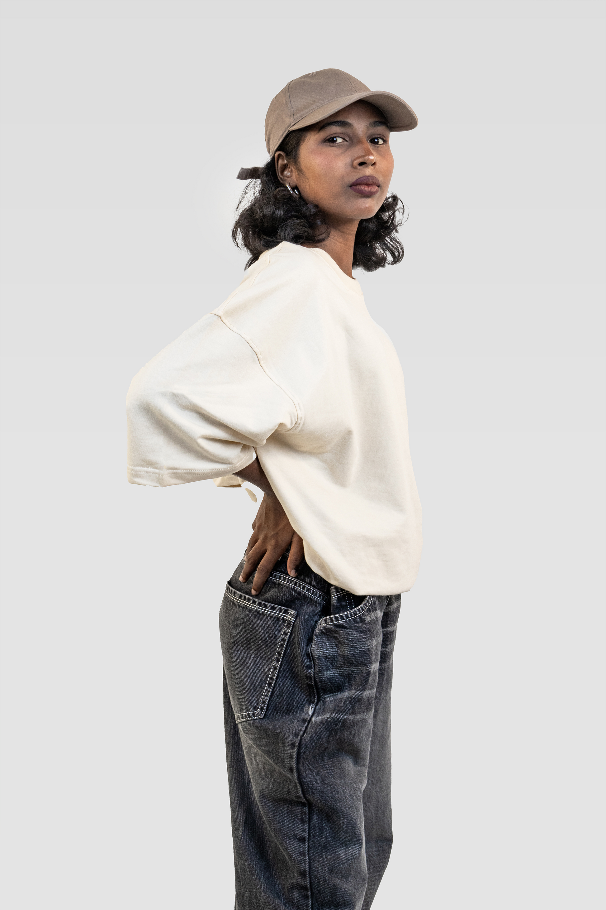 Side view of model wearing Harper Slein oversized t-shirt in OG Off White paired with denim jeans and a beige cap on a light gray background