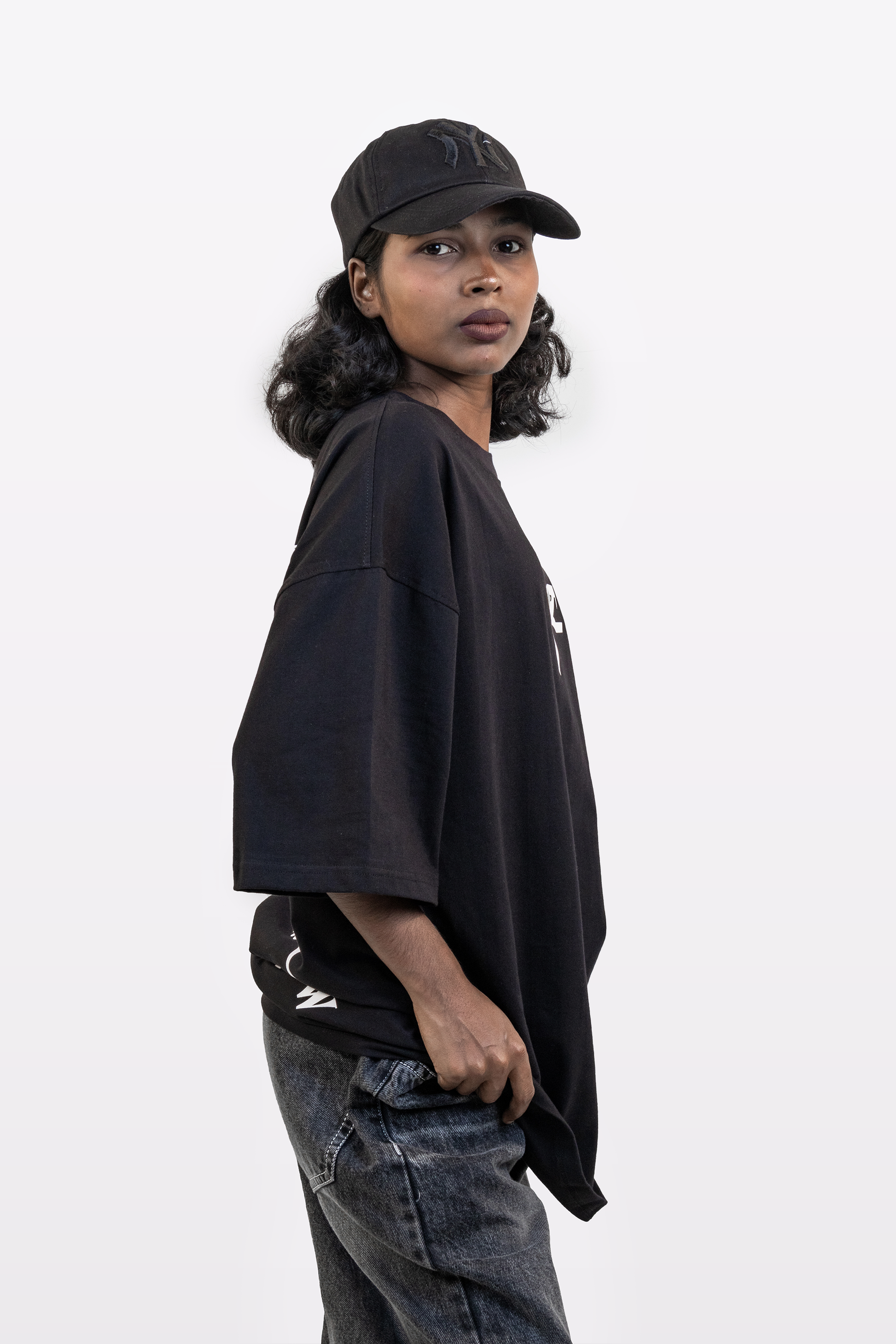 Side view of a model wearing the Harper Slein Urban Crew Collection black oversized T-shirt with snake logo, paired with a black cap and dark denim jeans, on a white background.
