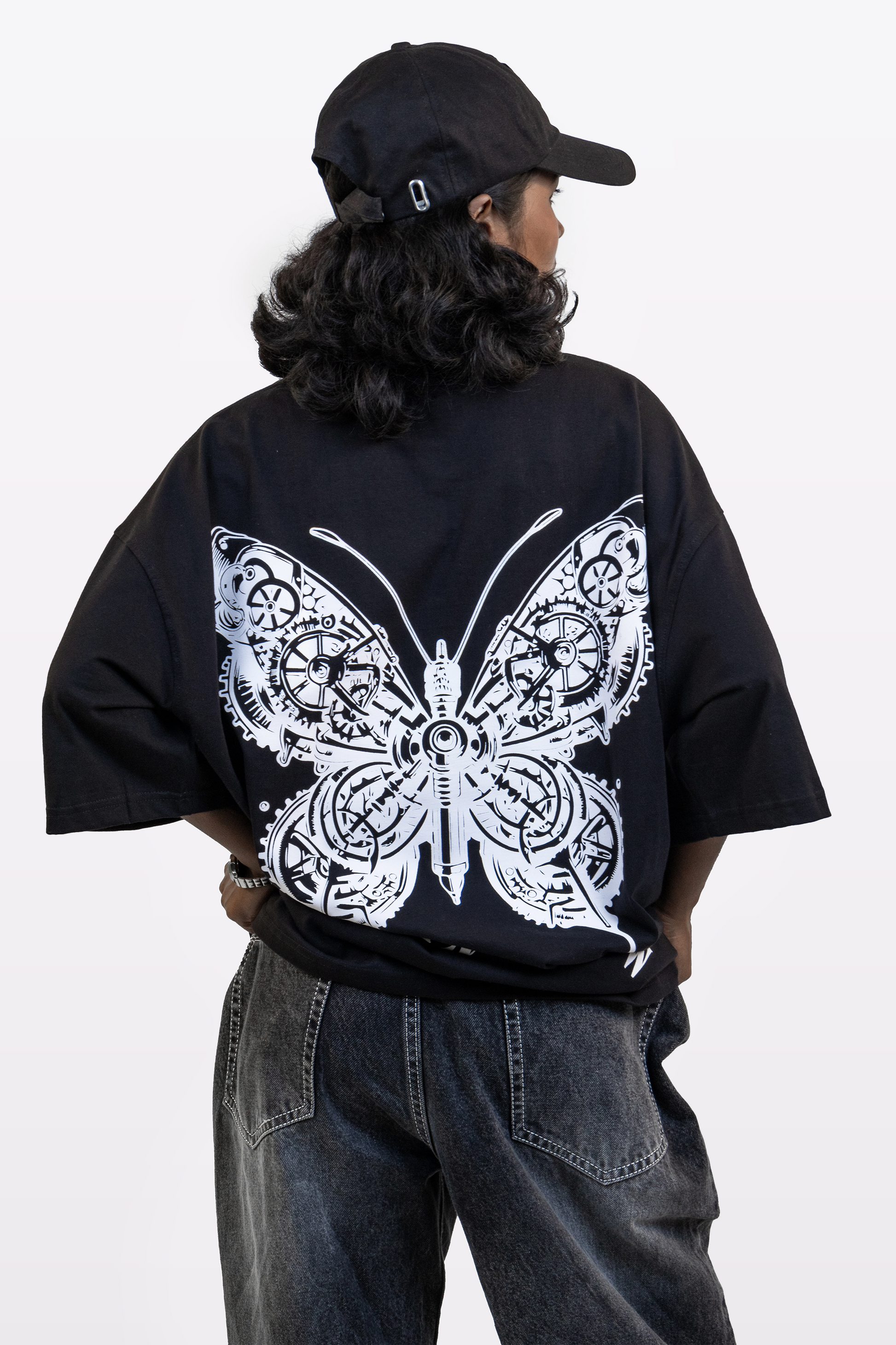 Back view of a model wearing the Harper Slein Urban Crew Collection black oversized T-shirt featuring a large white butterfly graphic, paired with a black cap and dark denim jeans, on a white background.