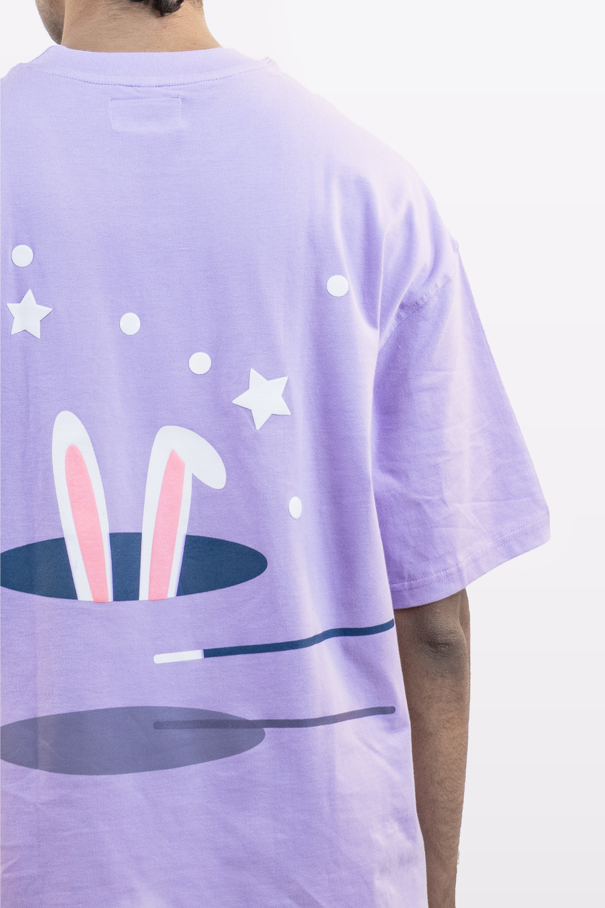 Close-up back view of a model wearing a lavender oversized t-shirt featuring a playful bunny ears and stars graphic design, against a plain white background.
