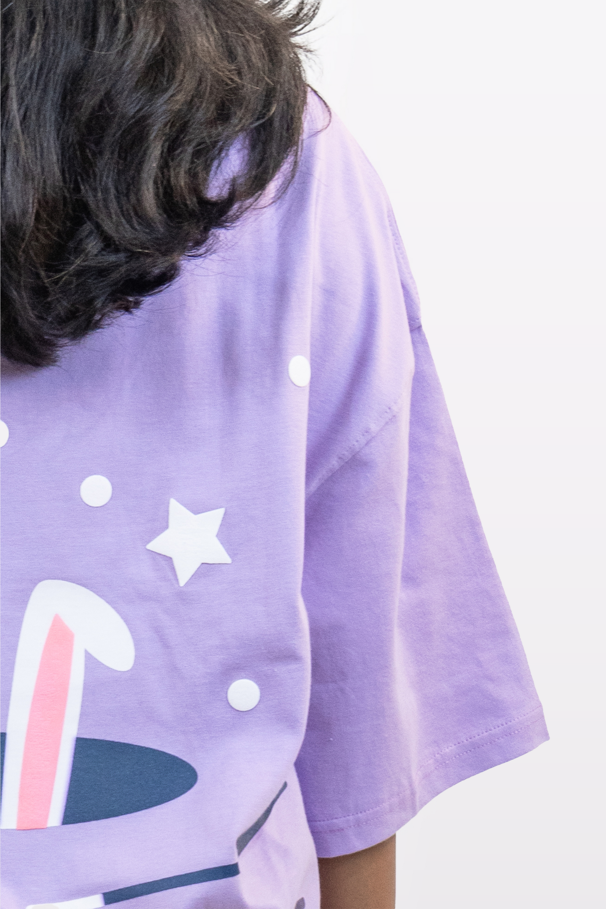 Close-up of a lavender t-shirt featuring a bunny ears and stars graphic on the back, worn by a model with long wavy hair, against a plain white background.
