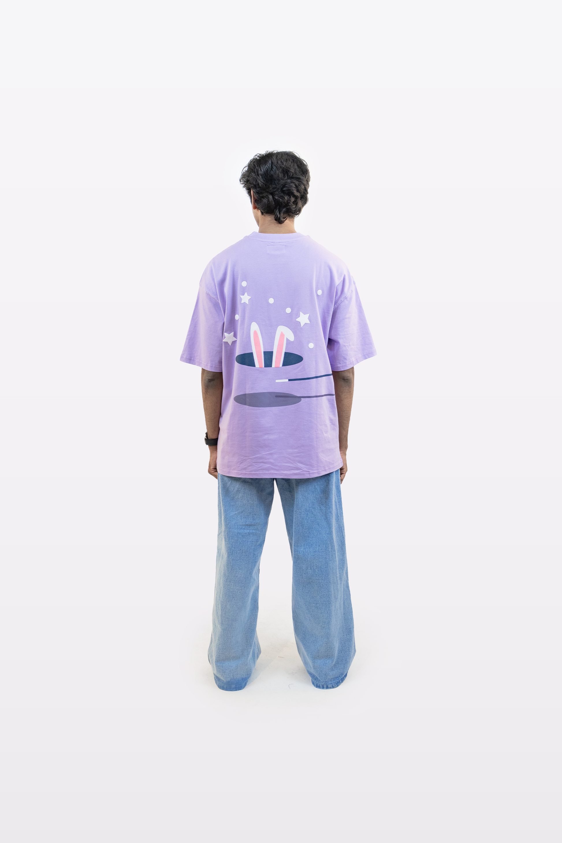Back view of a model wearing a lavender oversized t-shirt featuring a whimsical magician’s hat and bunny ears graphic, paired with light blue jeans, on a plain white background.