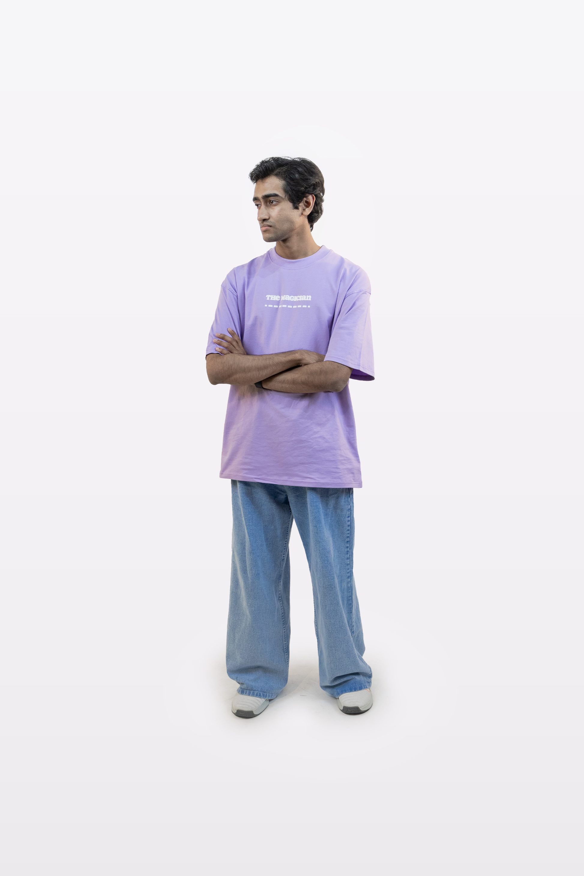Model standing with arms crossed, wearing a lavender oversized t-shirt with white front text and light blue jeans, on a plain white background.
