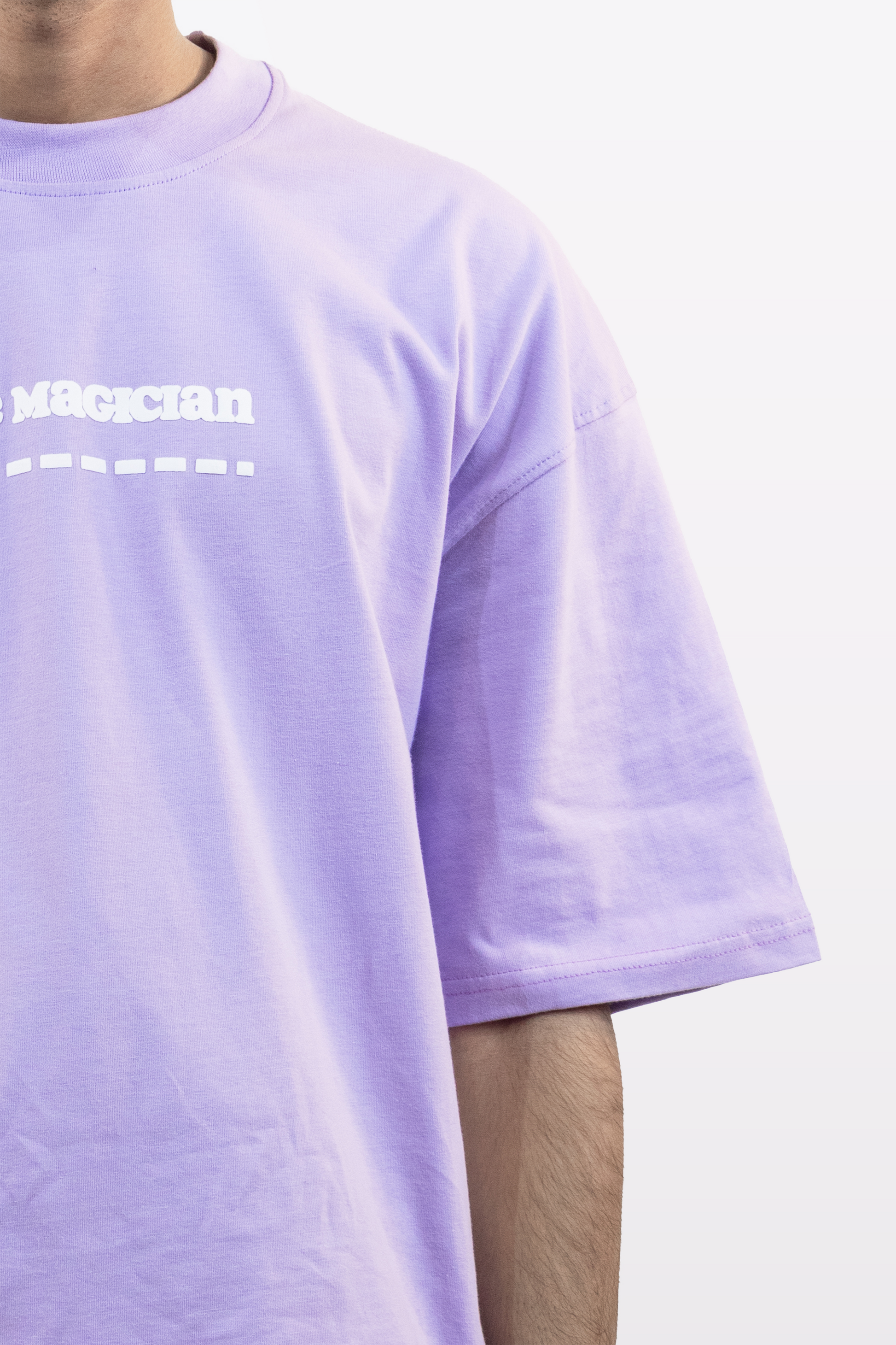 Close-up front view of a lavender oversized t-shirt with white ‘Magician’ text and design, worn by a model, against a plain white background.