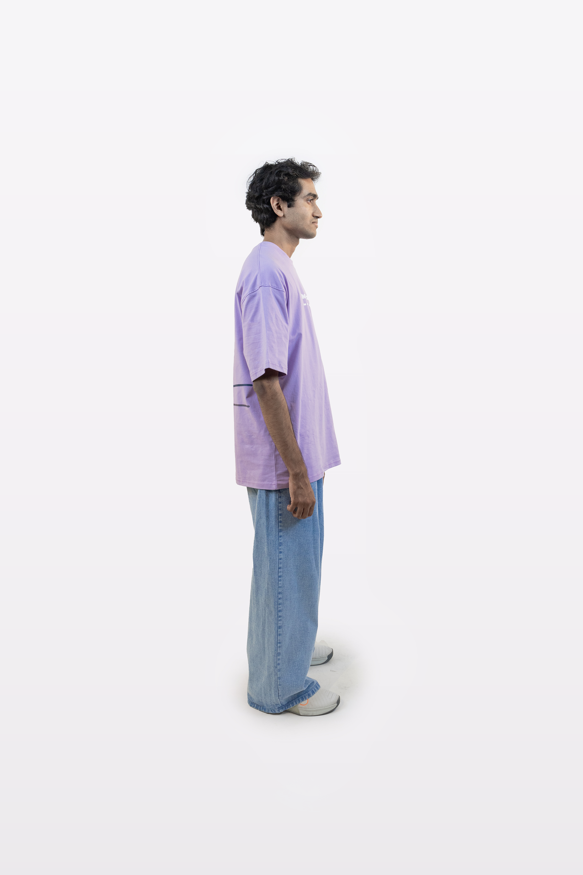 Side profile of a model wearing a lavender oversized t-shirt with white graphic text, paired with light blue jeans, on a plain white background.