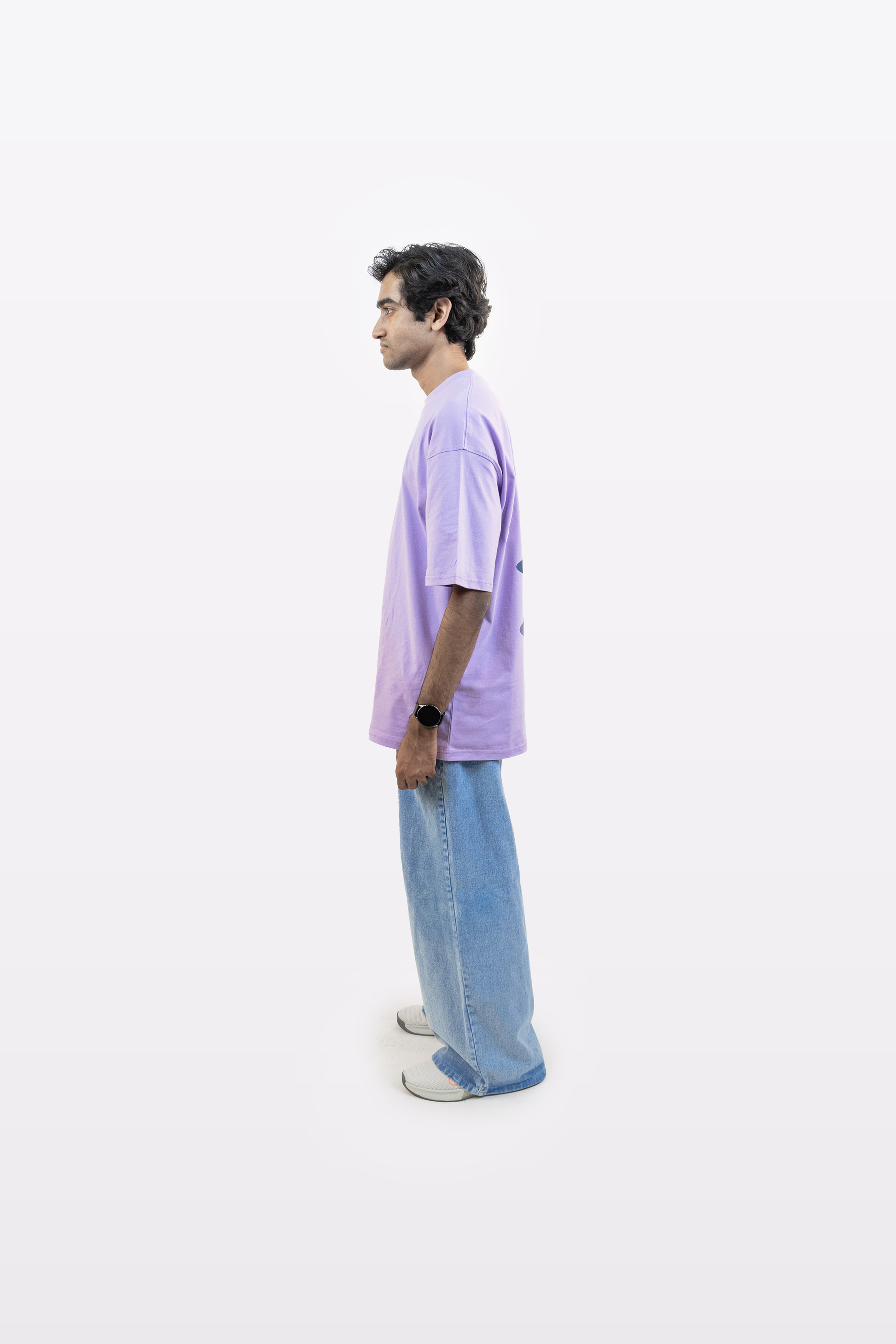 Right-side view of a model wearing a lavender oversized t-shirt with subtle front print and hidden back graphic, paired with light blue jeans, on a plain white background.