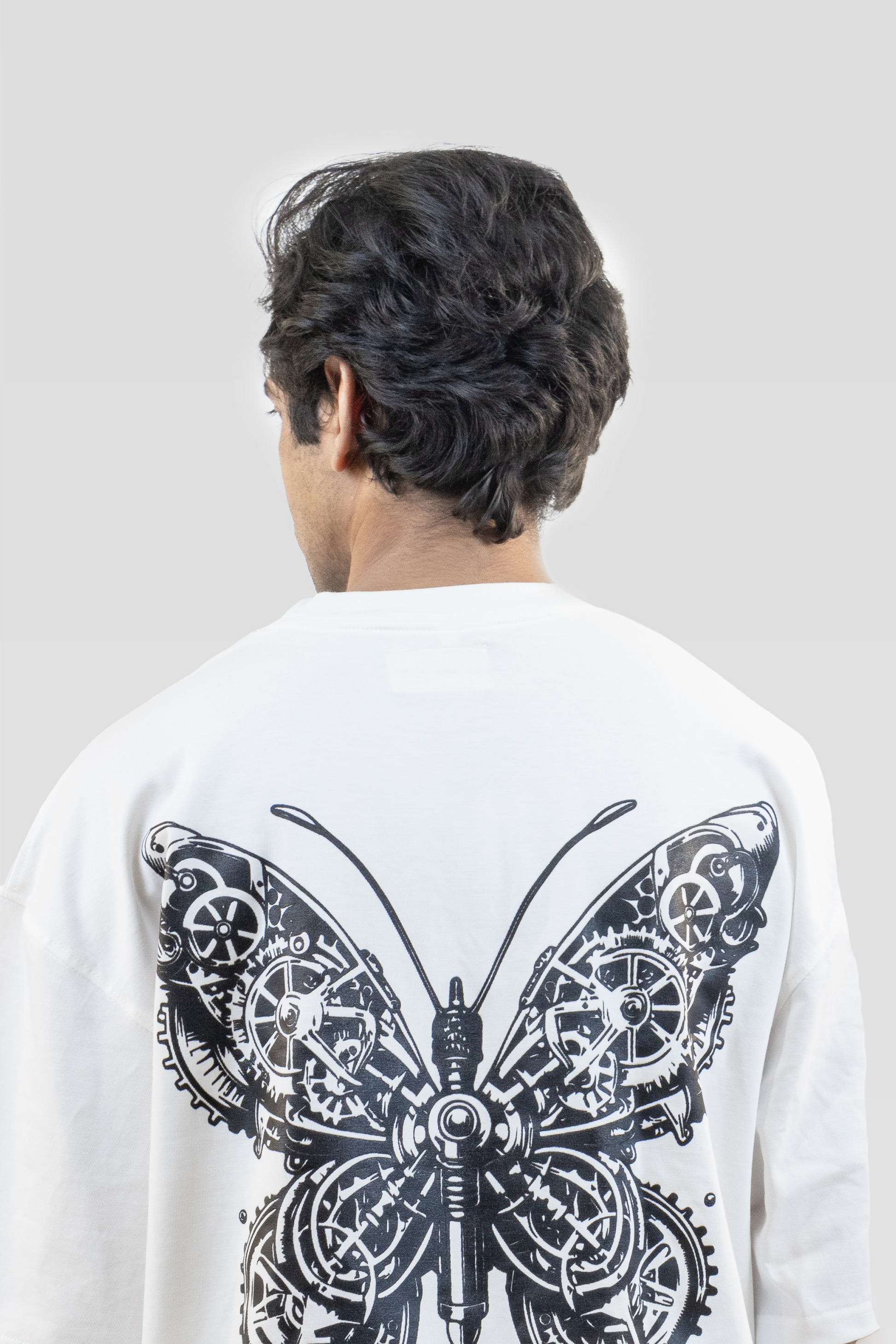 Close-up of the back of a white sweatshirt featuring a detailed black mechanical butterfly graphic, worn by a person standing against a light grey background.