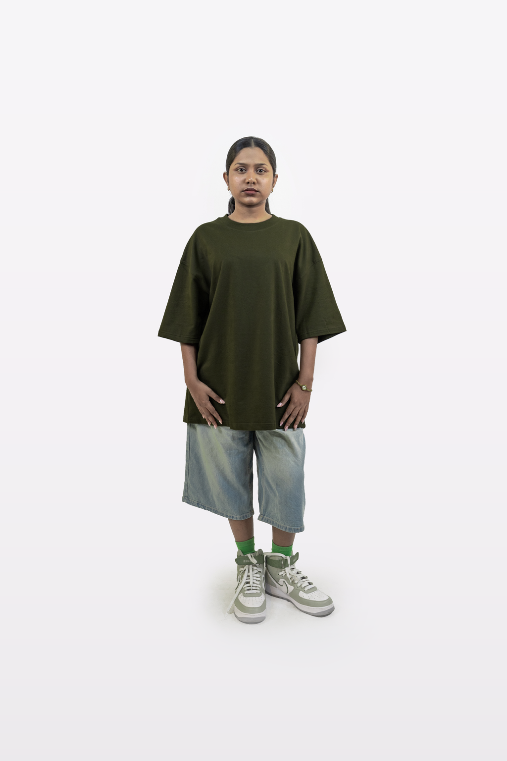 Full-body shot of a female model wearing the Harper Slein oversized t-shirt in Nori Seaweed Green, paired with knee-length denim shorts and green-accent sneakers, on a white background.