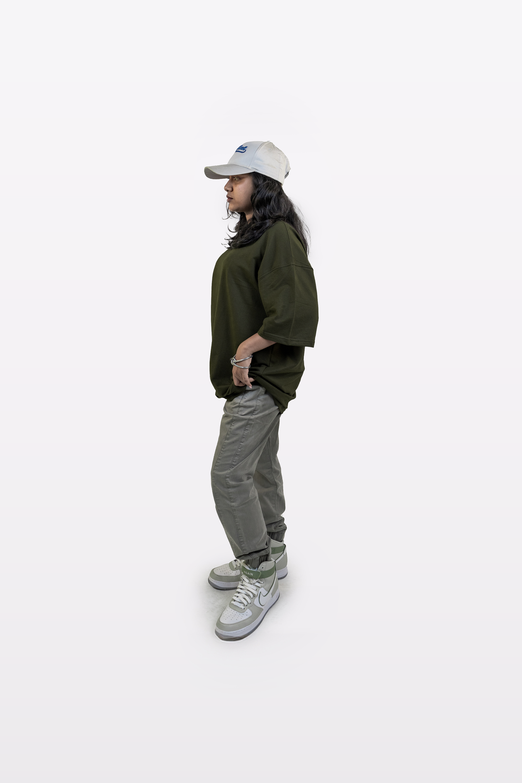 Side profile of a model wearing the Harper Slein oversized t-shirt in Nori Seaweed Green, paired with gray pants, white sneakers, and a white cap, standing against a white background.