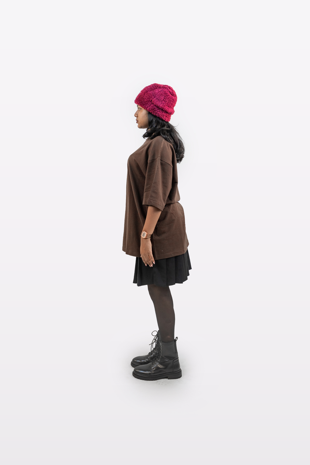Side profile of a female model wearing Harper Slein oversized t-shirt in Cacao styled with a black skirt, black boots, and a red beanie on a white background