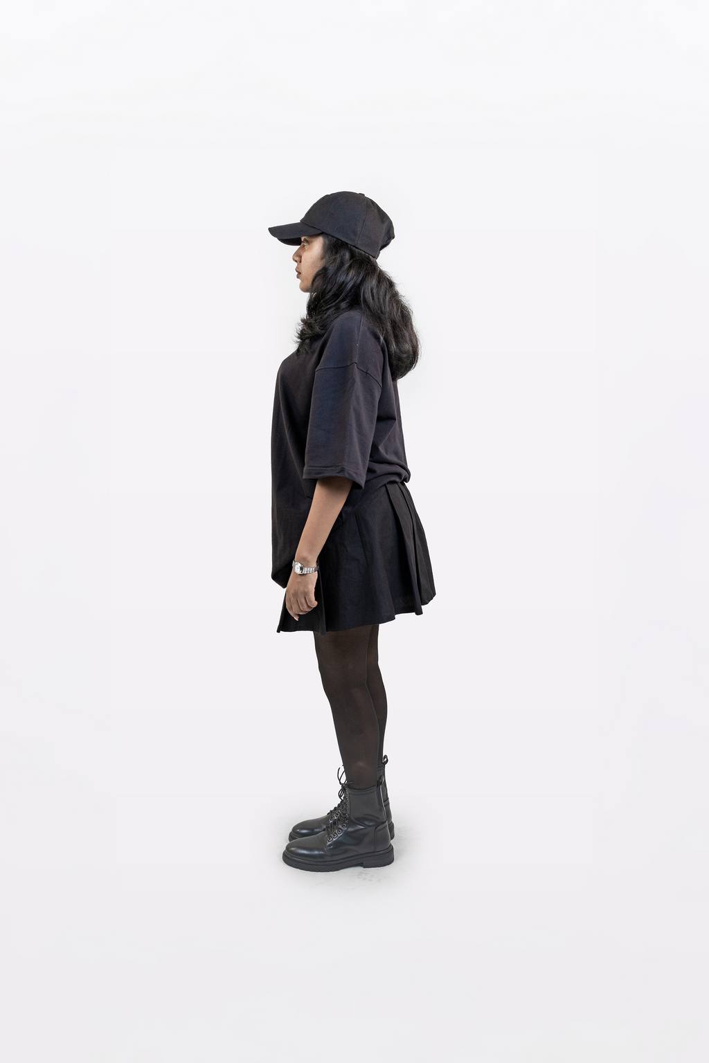 Upright side profile view of female model wearing Harper Slein oversized t-shirt in Classic Black, styled with black cap, skirt, tights, and boots on a white background