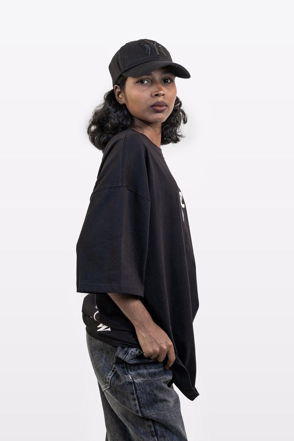 Side view of a model wearing the Harper Slein Urban Crew Collection black oversized T-shirt with snake logo, paired with a black cap and dark denim jeans, on a white background.