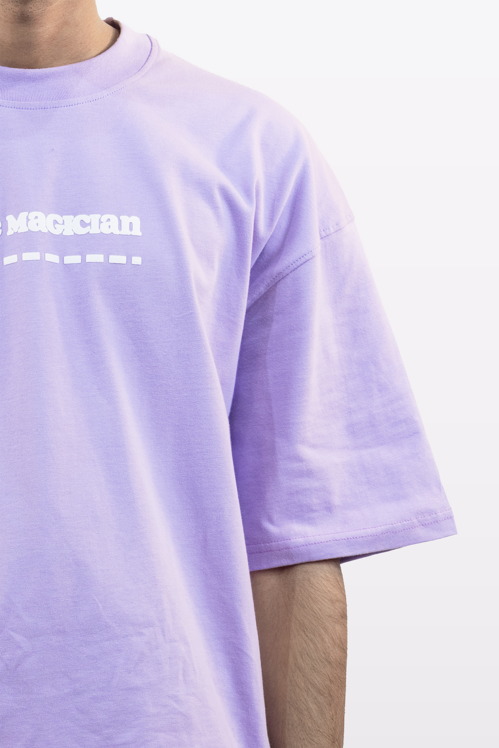 Close-up front view of a lavender oversized t-shirt with white ‘Magician’ text and design, worn by a model, against a plain white background.