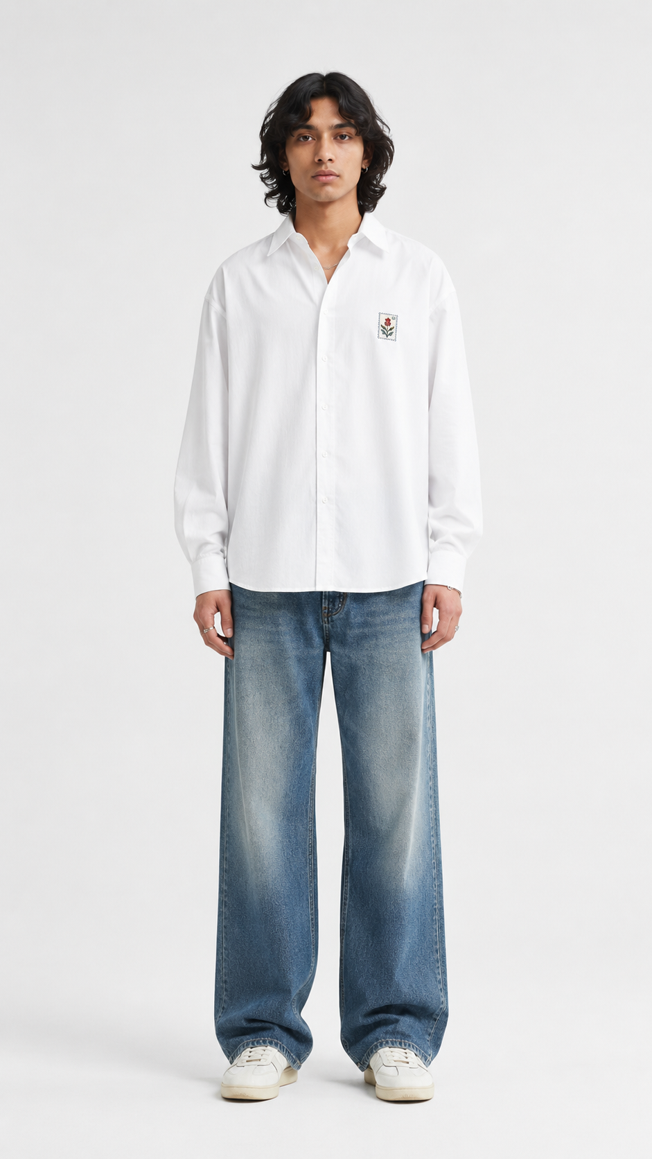 A model stands against a light gray studio background wearing a white long-sleeve button-up shirt with a small embroidered crest on the left chest, loose-fit blue jeans, and white sneakers.