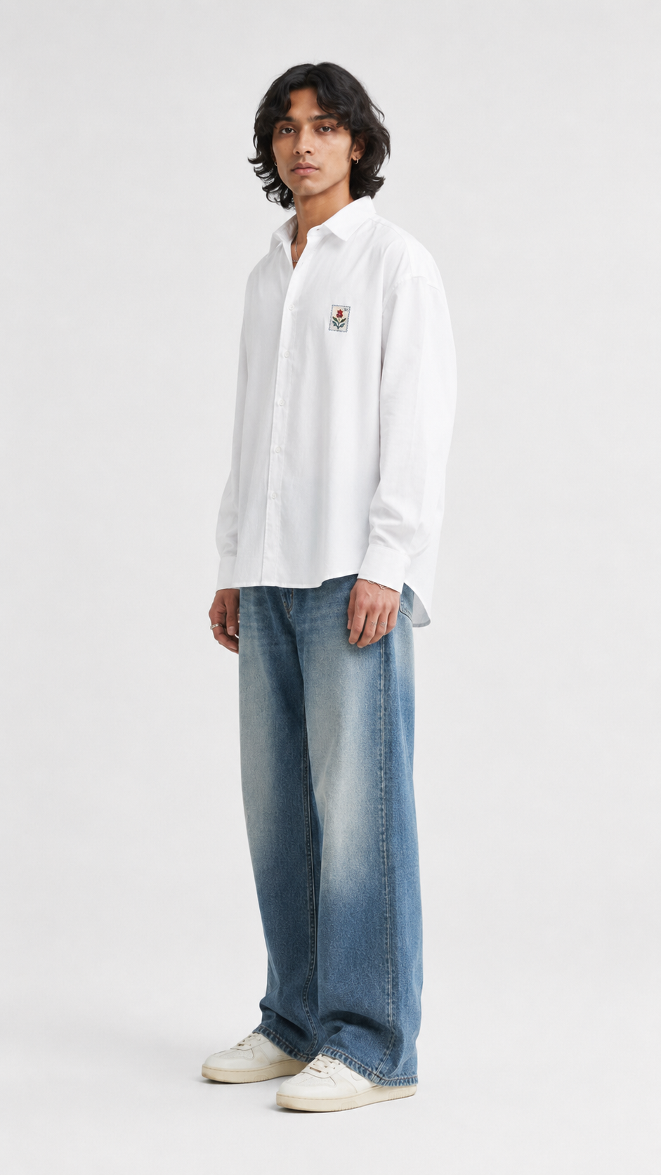 A man wearing a white long-sleeve button-up shirt with a small embroidered logo on the left chest, paired with wide-leg faded blue jeans and white sneakers, standing against a light gray studio background.