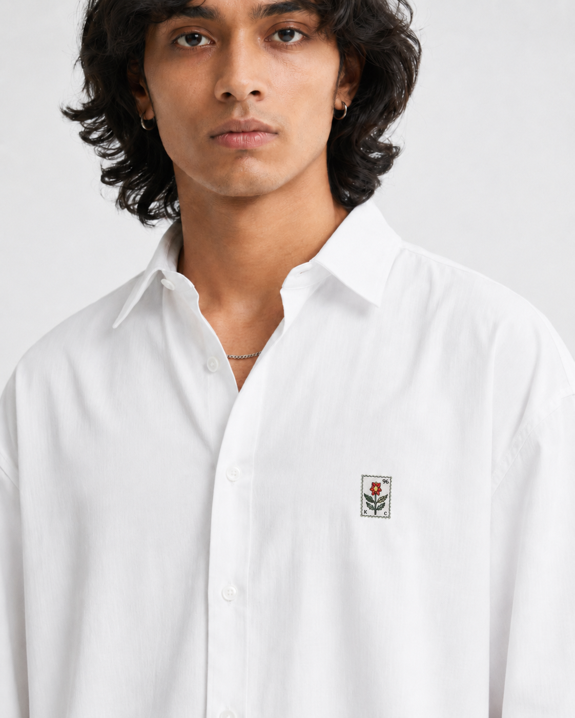 A close-up of a man wearing a white long-sleeve button-down shirt with a small embroidered shield logo on the left chest, set against a plain light gray background.