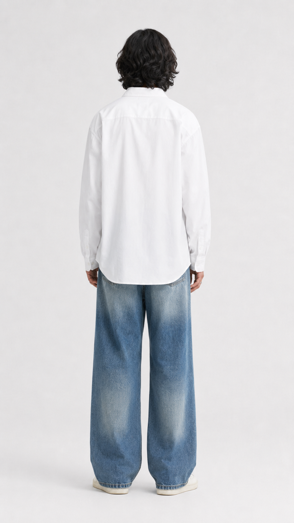 A model viewed from the back wearing a white long-sleeve button-up shirt and wide-leg faded blue jeans, standing against a light gray studio background.